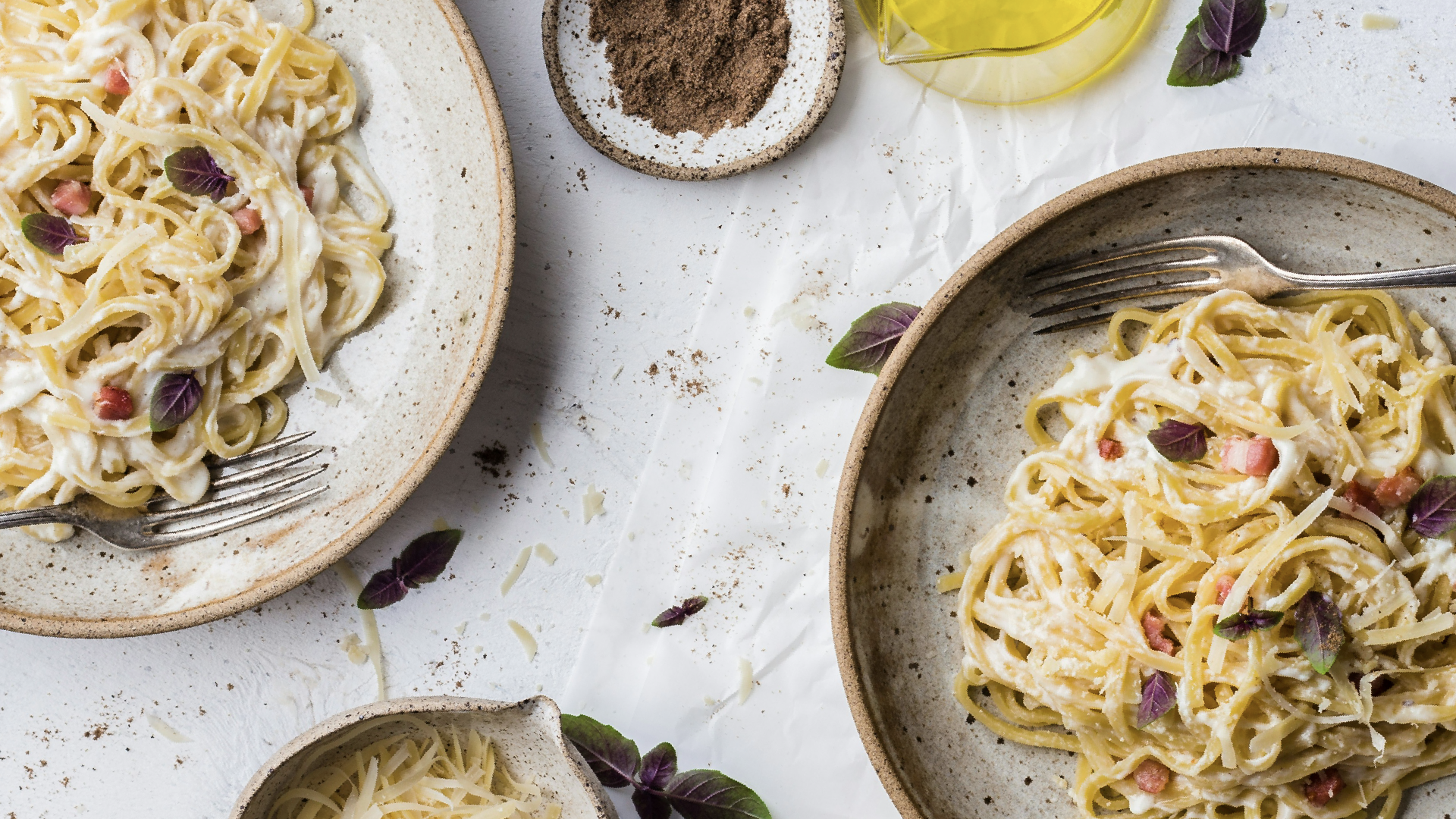 Creamy pasta dishes from top restaurants in Scarsdale, garnished with basil and pancetta, alongside bowls of grated cheese and ground pepper.