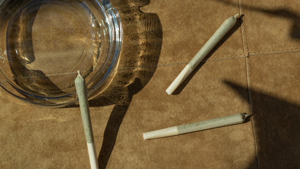 Three infused prerolls placed on a brown suede surface beside a clear glass ashtray, with shadows cast by the sunlight.