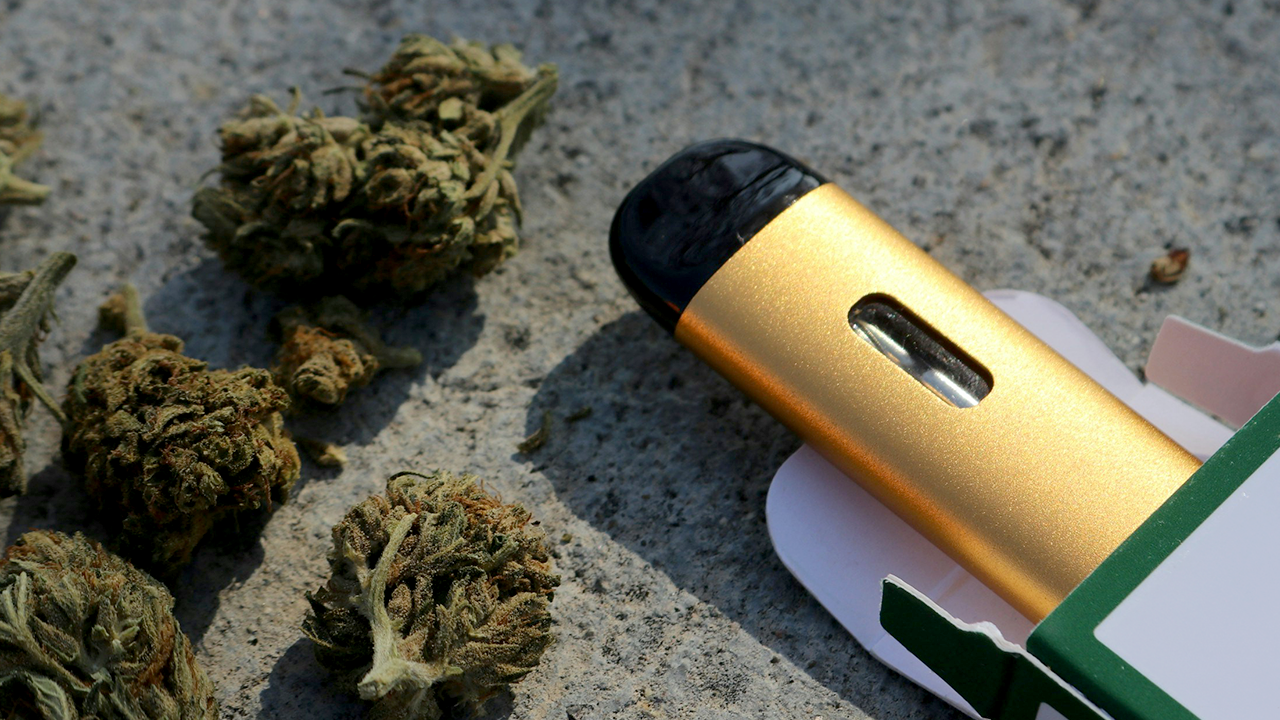 Gold all-in-one disposable vape pen with cannabis flower on a stone surface.